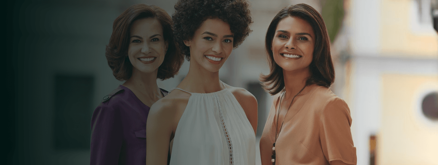 São três mulheres sorrindo, a direita é branca com blusa roxa, a do meio negra de cabelos cacheados e a esquerda parda de blusa laranja