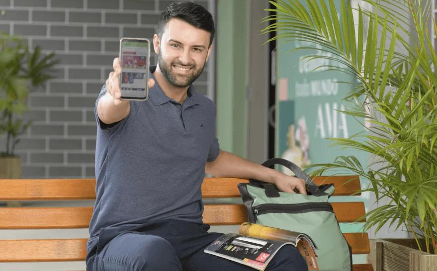 Homem branco, cabelo curto preto e de barba curta, segurando um celular com o aplicativo do Revendedor O Boticário aberto.