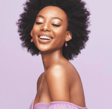 Mulher negra de cabelos cacheados, com maquiagem no rosto, vestindo uma blusa rosa “ombro a ombro”, brincos e sorrindo de olhos fechados