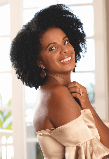 Mulher negra de cabelos cacheados, com maquiagem no rosto, vestindo uma blusa bege “ombro a ombro”, brincos e sorrindo